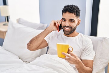 Young hispanic man talking on smartphone drinking coffee sitting on bed at bedroom