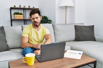 Young man with beard using laptop at home winking looking at the camera with sexy expression,...