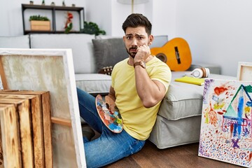Young man with beard painting canvas at home looking stressed and nervous with hands on mouth biting nails. anxiety problem.