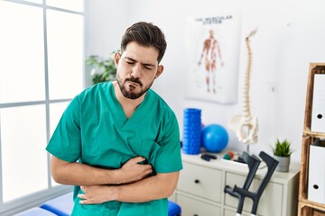 Young man with beard working at pain recovery clinic with hand on stomach because nausea, painful disease feeling unwell. ache concept.