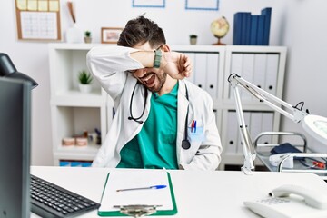 Young man with beard wearing doctor uniform and stethoscope at the clinic covering eyes with arm smiling cheerful and funny. blind concept.