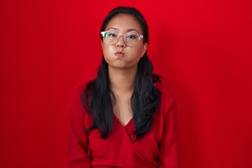 Asian young woman standing over red background puffing cheeks with funny face. mouth inflated with air, crazy expression. - obrazy, fototapety, plakaty