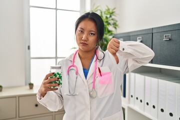 Young asian doctor woman holding pills with angry face, negative sign showing dislike with thumbs...