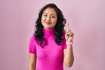Young asian woman standing over pink background showing and pointing up with finger number one while smiling confident and happy.