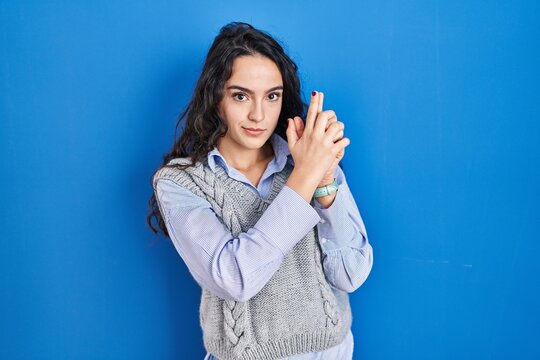 Young brunette woman standing over blue background holding symbolic gun with hand gesture, playing killing shooting weapons, angry face