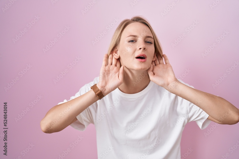 Poster young caucasian woman standing over pink background trying to hear both hands on ear gesture, curiou