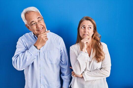 Middle age hispanic couple standing over blue background thinking worried about a question, concerned and nervous with hand on chin
