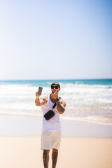 Chico joven musculado posando con cinta en la cabeza en la playa con el smartphone en la mano