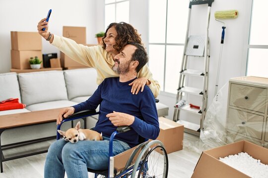 Middle age hispanic couple smiling happy. Man sitting on wheelchair with dog on his legs and woman making selfie by the smartphone at new home.