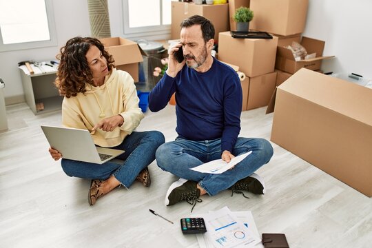 Middle age hispanic couple controlling family economy using laptop. Sitting on the floor talking on the smartphone at new home.