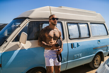 Chico joven tatuado y musculoso posando delante de una furgoneta celeste y blanca en zona de campers