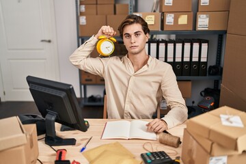 Young man working at small business ecommerce holding alarm clock thinking attitude and sober...