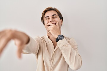 Young man standing over isolated background laughing at you, pointing finger to the camera with hand over body, shame expression