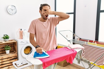 Young hispanic man ironing clothes at home peeking in shock covering face and eyes with hand, looking through fingers with embarrassed expression.