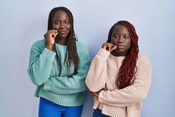 Two african woman standing over blue background mouth and lips shut as zip with fingers. secret and...