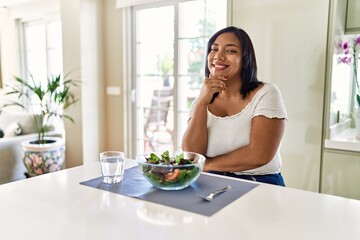 Obraz na płótnie Canvas Young hispanic woman eating healthy salad at home looking confident at the camera smiling with crossed arms and hand raised on chin. thinking positive.