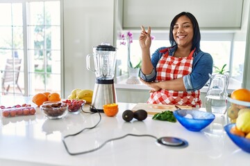 Young hispanic woman making healthy smoothie smiling with happy face winking at the camera doing...