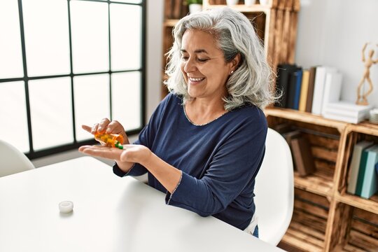 Middle age grey-haired woman taking pills sitting on the table at home.