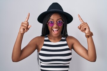 Young african american with braids wearing hat and sunglasses smiling amazed and surprised and pointing up with fingers and raised arms.