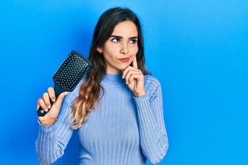 Young hispanic girl holding hairbrush serious face thinking about question with hand on chin,...