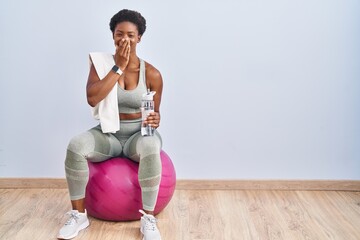 African american woman wearing sportswear sitting on pilates ball laughing and embarrassed giggle...