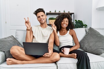 Young interracial couple using laptop at home sitting on the sofa smiling with happy face winking at the camera doing victory sign with fingers. number two.