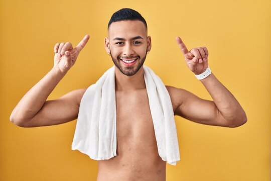 Young hispanic man standing shirtless with towel smiling amazed and surprised and pointing up with fingers and raised arms.