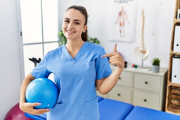 Young physiotherapist woman holding pilates ball at medical clinic pointing finger to one self smiling happy and proud