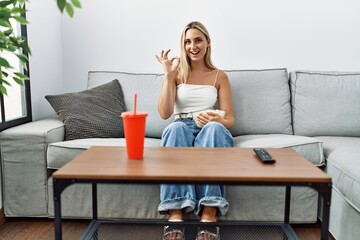 Young blonde woman eating popcorn sitting on the sofa doing ok sign with fingers, smiling friendly gesturing excellent symbol
