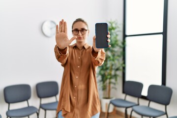Young blonde woman holding smartphone showing blank screen with open hand doing stop sign with serious and confident expression, defense gesture