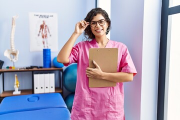 Young latin woman wearing physiotherapist uniform holding clipboard at clinic