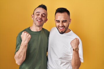 Homosexual couple standing over yellow background very happy and excited doing winner gesture with arms raised, smiling and screaming for success. celebration concept.