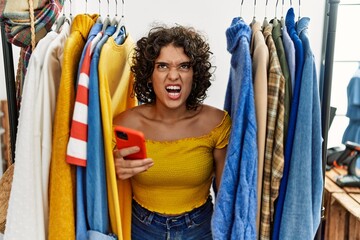 Young hispanic woman searching clothes on clothing rack using smartphone angry and mad screaming...