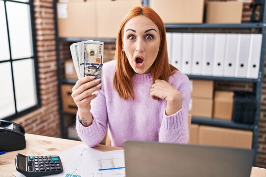 Young woman working at small business ecommerce holding money scared and amazed with open mouth for surprise, disbelief face