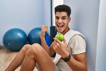 Young hispanic man wearing sportswear eating healthy apple pointing thumb up to the side smiling happy with open mouth