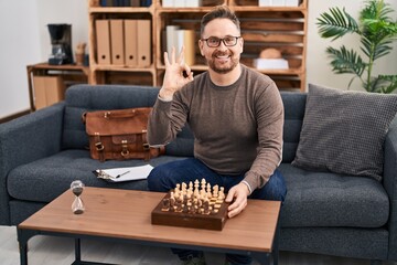 Middle age caucasian man playing chess sitting on the sofa doing ok sign with fingers, smiling friendly gesturing excellent symbol