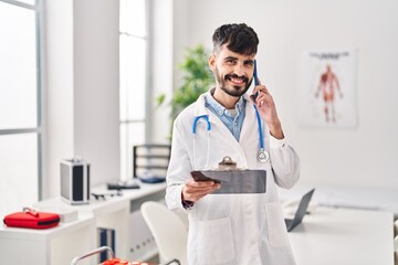 Young hispanic man wearing doctor uniform talking on smartphone reading medical report at clinic