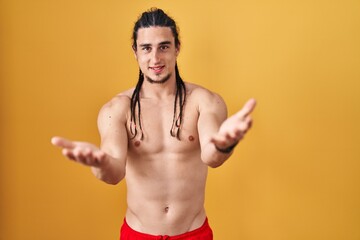 Fototapeta premium Hispanic man with long hair standing shirtless over yellow background smiling cheerful offering hands giving assistance and acceptance.