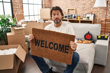 Handsome middle age man holding welcome doormat at new home clueless and confused expression. doubt concept.