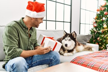 Young hispanic man holding gift sitting on sofa with dog by christmas tree at home
