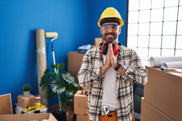 Young hispanic man with beard working at home renovation praying with hands together asking for forgiveness smiling confident.