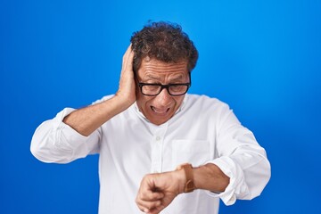 Middle age hispanic man standing over blue background looking at the watch time worried, afraid of...