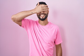 Hispanic young man standing over pink background smiling and laughing with hand on face covering eyes for surprise. blind concept.