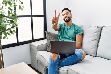 Young handsome man with beard using computer laptop sitting on the sofa at home smiling looking to...