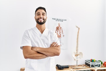 Young arab man wearing physiotherapist uniform standing with arms crossed gesture at clinic - obrazy, fototapety, plakaty
