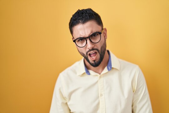 Hispanic young man wearing business clothes and glasses in shock face, looking skeptical and sarcastic, surprised with open mouth