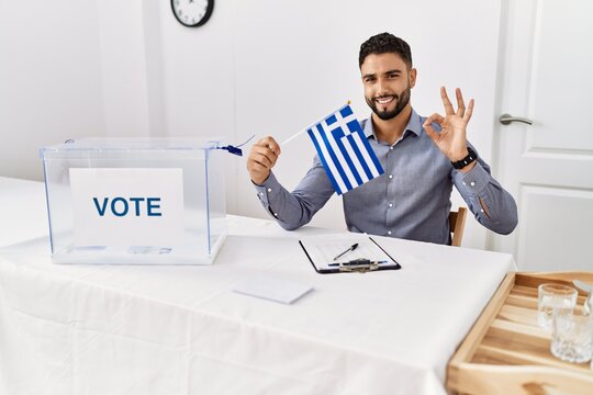 Young handsome man with beard at political campaign election holding greece flag doing ok sign with fingers, smiling friendly gesturing excellent symbol