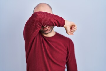 Young bald man with beard standing over white background wearing glasses covering eyes with arm smiling cheerful and funny. blind concept.