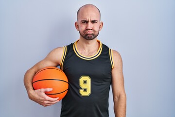 Young bald man with beard wearing basketball uniform holding ball puffing cheeks with funny face. mouth inflated with air, crazy expression.