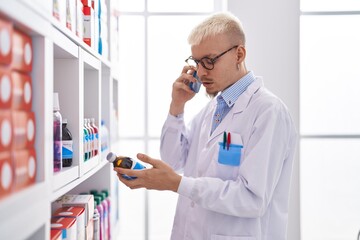 Young caucasian man pharmacist talking on smartphone holding medicine bottle at pharmacy